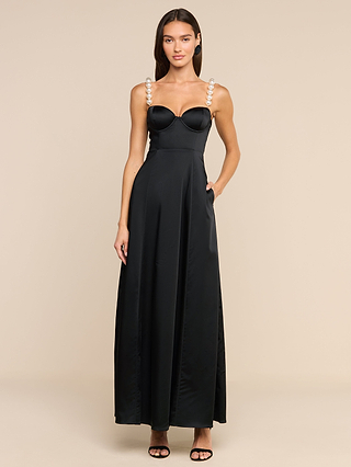 A woman with long brown hair stands against a plain beige background wearing the Sutton Maxi Dress, a long black satin evening gown featuring simulated pearl straps. She pairs the look with black high-heeled sandals and keeps her hands relaxed at her sides.