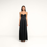 A woman stands facing forward against a plain white background, wearing the Sutton Maxi Dress—a black satin evening gown featuring a fitted bodice and flared skirt. She has long brown hair and pairs the dress with black open-toe heels.