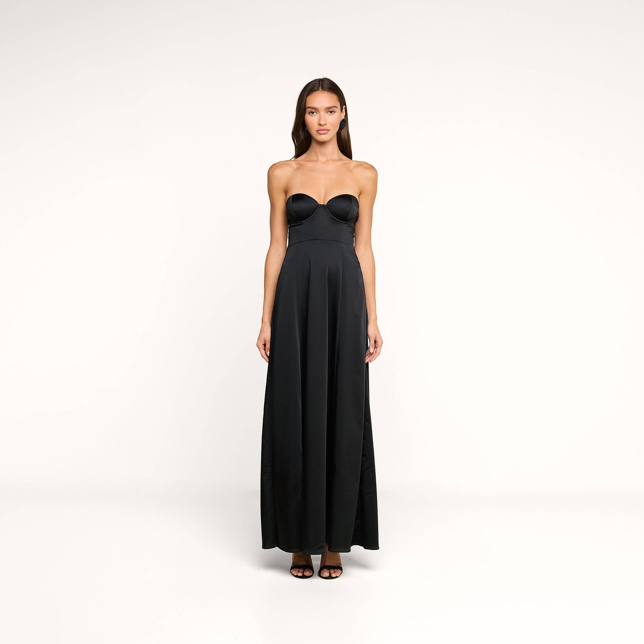 A woman stands facing forward against a plain white background, wearing the Sutton Maxi Dress—a black satin evening gown featuring a fitted bodice and flared skirt. She has long brown hair and pairs the dress with black open-toe heels.