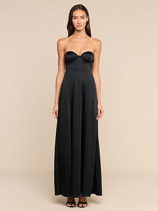 A woman stands facing forward, wearing the Sutton Maxi Dress—a strapless, floor-length black satin gown with a fitted bodice and flared skirt. She has long brown hair and is wearing black open-toe heels, set against a plain beige background.