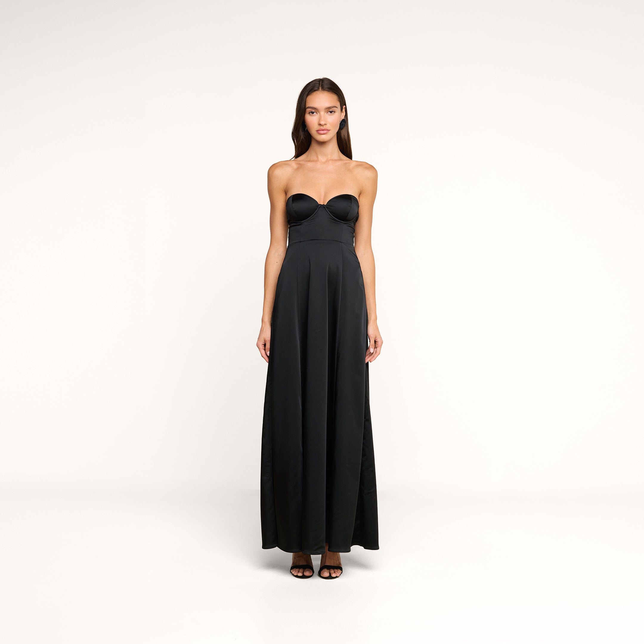 A woman stands facing forward against a plain white background, wearing the Sutton Maxi Dress—a black satin evening gown featuring a fitted bodice and flared skirt. She has long brown hair and pairs the dress with black open-toe heels.