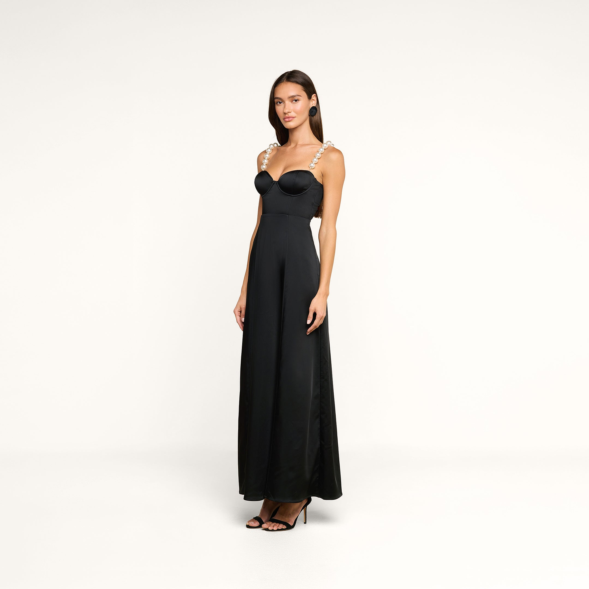 A woman stands in a studio wearing the Sutton Maxi Dress, a long black satin evening gown with pearl straps, paired with black high-heeled shoes. She faces forward with her head slightly turned against a plain white background.