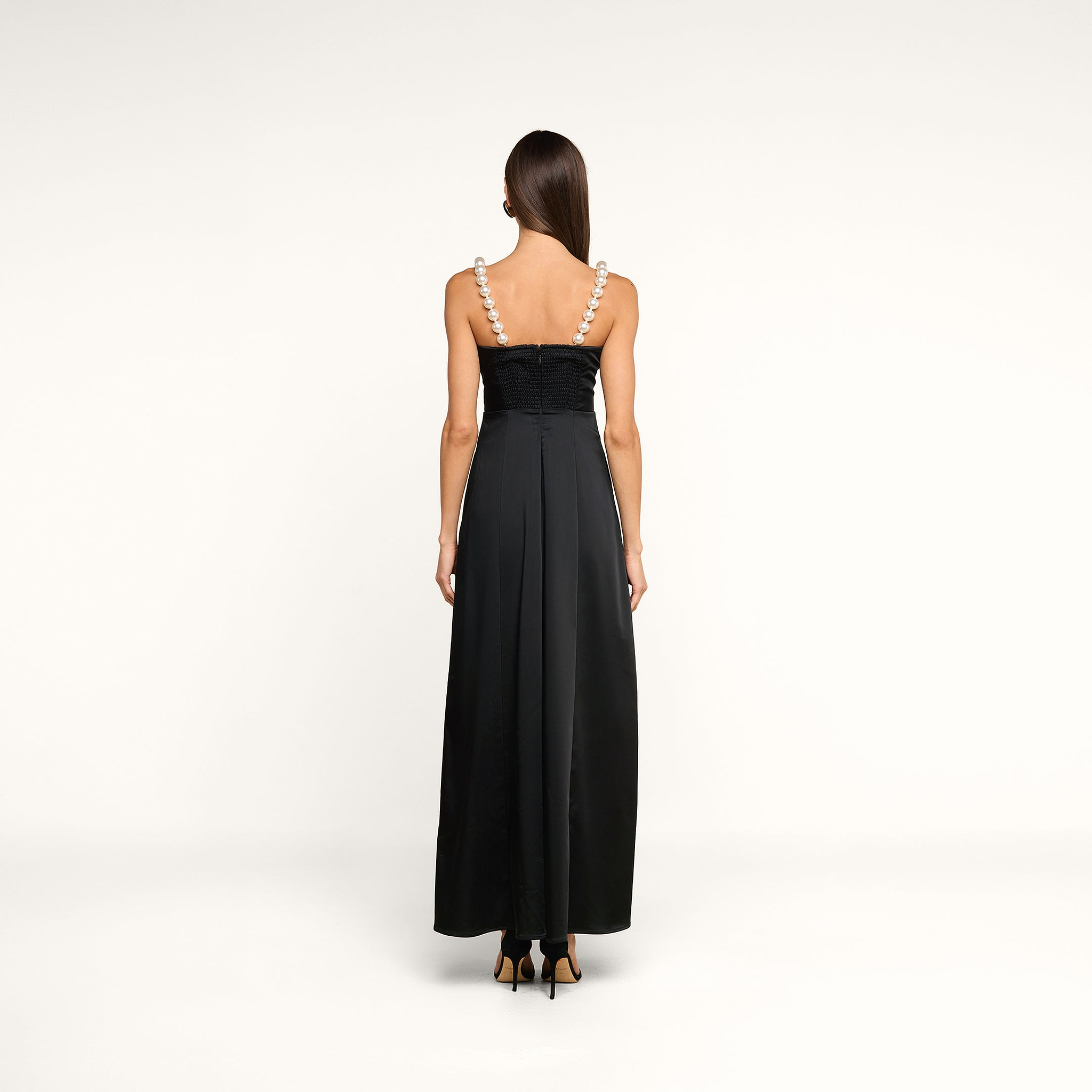 A woman stands with her back to the camera, wearing the Sutton Maxi Dress—a long black satin evening gown with wide pearl straps—paired with black high-heeled shoes, set against a plain white background.