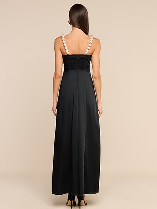 A woman with straight brown hair, wearing the Sutton Maxi Dress—a long black satin evening gown featuring large simulated pearl straps—stands with her back to the camera against a beige background. She completes her look with black high heels.
