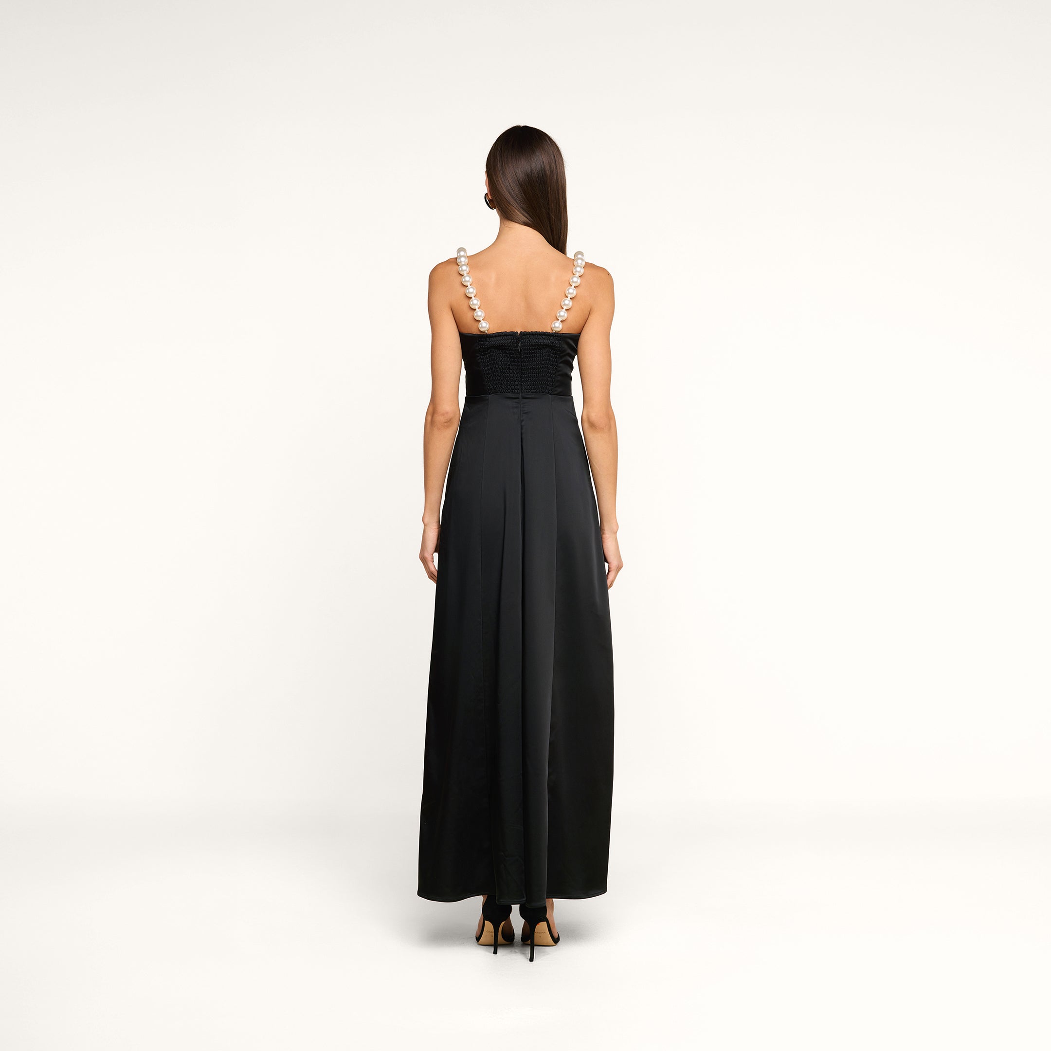 A woman stands with her back to the camera, wearing the Sutton Maxi Dress—a long black satin evening gown with wide pearl straps—paired with black high-heeled shoes, set against a plain white background.