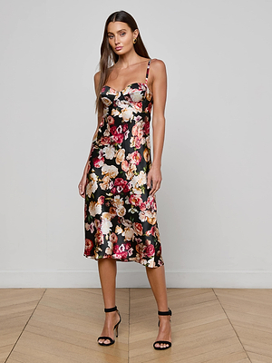 A woman with long brown hair stands on a wooden floor against a plain light gray background, wearing the L'AGENCE Solay Silk Slip Dress, a black, sleeveless rose-print dress with thin straps, and black high-heeled sandals.