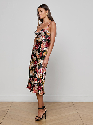 A woman stands on a wooden floor wearing the Solay Silk Slip Dress by L’AGENCE, featuring a black base with a vibrant floral print and spaghetti straps. She pairs it with black high-heeled sandals while posing sideways against a white wall.