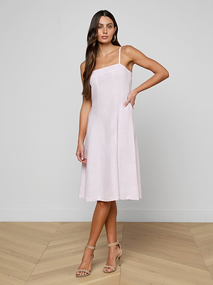 A woman in the Mitzy Linen Midi Dress, a sleeveless light pink linen day dress with adjustable straps, stands on a wooden floor against a plain light gray wall. She wears beige heels and poses with one hand on her hip, her long brown hair flowing.