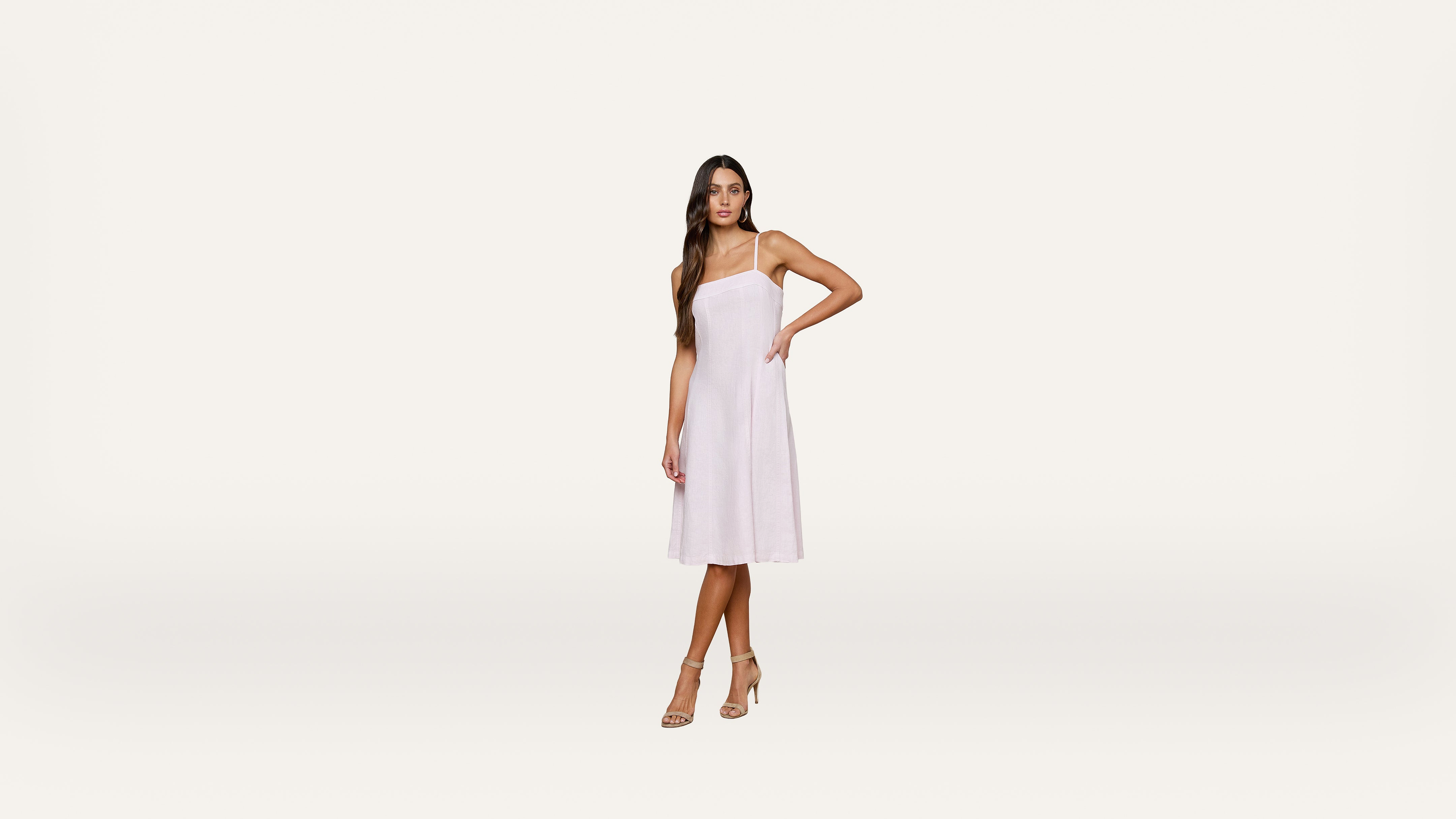 Woman wearing a sleeveless, light pink midi dress with spaghetti straps and nude heels, standing against a plain white background.