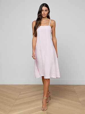 A woman with long brown hair wears the Mitzy Linen Midi Dress, a light pink fit-and-flare style with adjustable straps, and beige high-heeled sandals, standing on a light wood floor against a plain light gray wall.