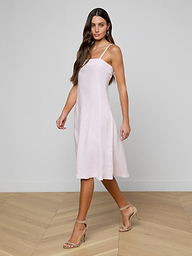 A woman with long brown hair walks indoors on a light wood floor, wearing the Mitzy Linen Midi Dress—a light pink, sleeveless fit-and-flare style with adjustable straps—paired with beige high-heeled sandals against a plain white wall.