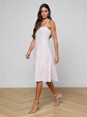 A woman with long brown hair walks indoors on a light wood floor, wearing the Mitzy Linen Midi Dress—a light pink, sleeveless fit-and-flare style with adjustable straps—paired with beige high-heeled sandals against a plain white wall.