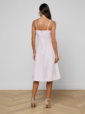 A woman stands facing away, wearing the pale pink Mitzy Linen Midi Dress with adjustable straps and a back zipper. The sleeveless, fit-and-flare dress falls to her knees and is paired with beige high-heeled sandals on a wooden floor against a plain wall.
