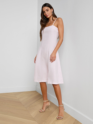A woman stands in a corner of a room wearing the Mitzy Linen Midi Dress, a light pink fit-and-flare style with adjustable straps, paired with beige high-heeled sandals. She poses with one hand by her side and the other gently touching her dress.