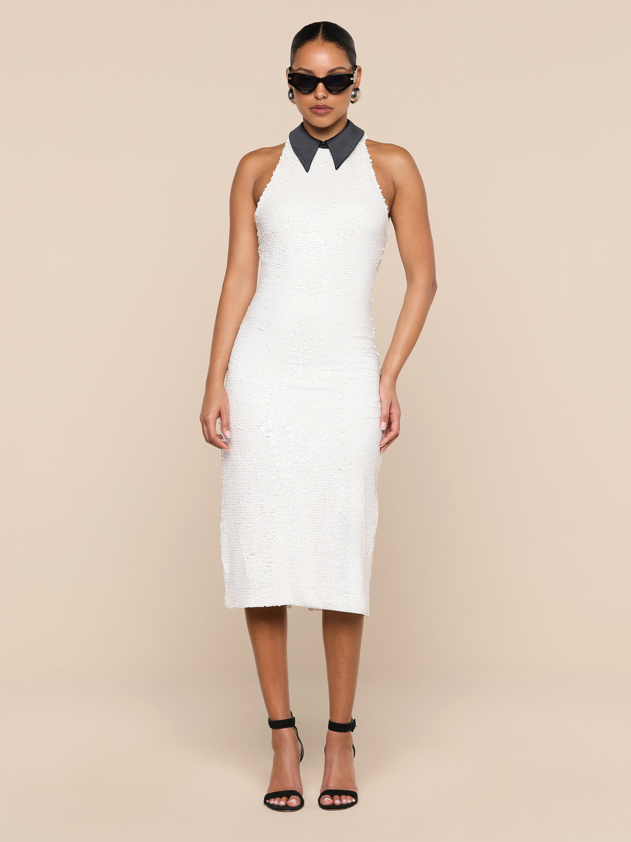 A woman wearing the Lilith Sequin Dress—a sleeveless, knee-length fitted party dress with a black collar—paired with black high heels and sunglasses, stands against a plain white background.