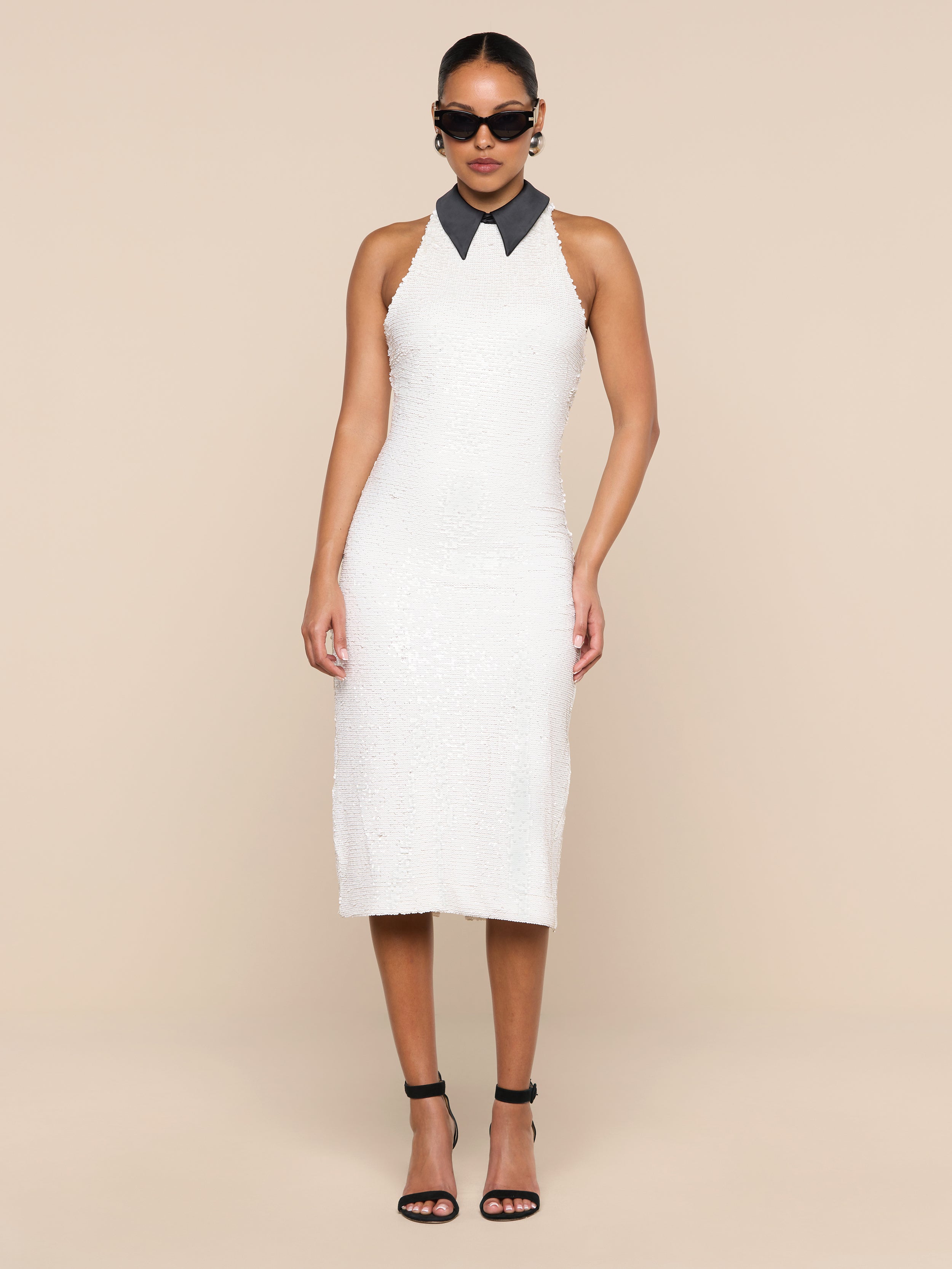 A woman wearing the Lilith Sequin Dress—a sleeveless, knee-length fitted party dress with a black collar—paired with black high heels and sunglasses, stands against a plain white background.