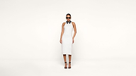 A woman wearing a sleeveless white dress, black strappy heels, and dark sunglasses stands against a plain white background.
