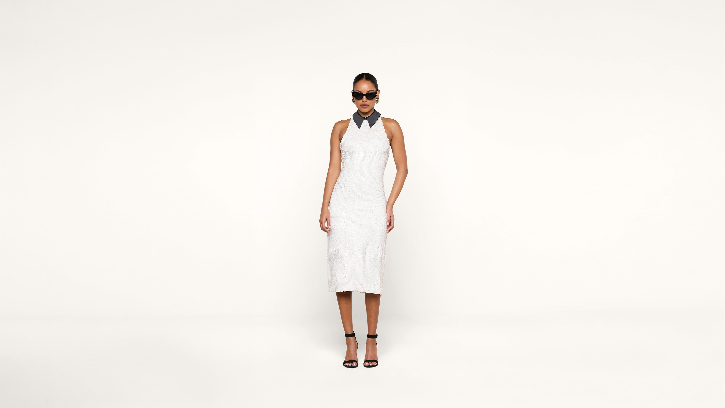 A woman wearing a sleeveless white dress, black strappy heels, and dark sunglasses stands against a plain white background.