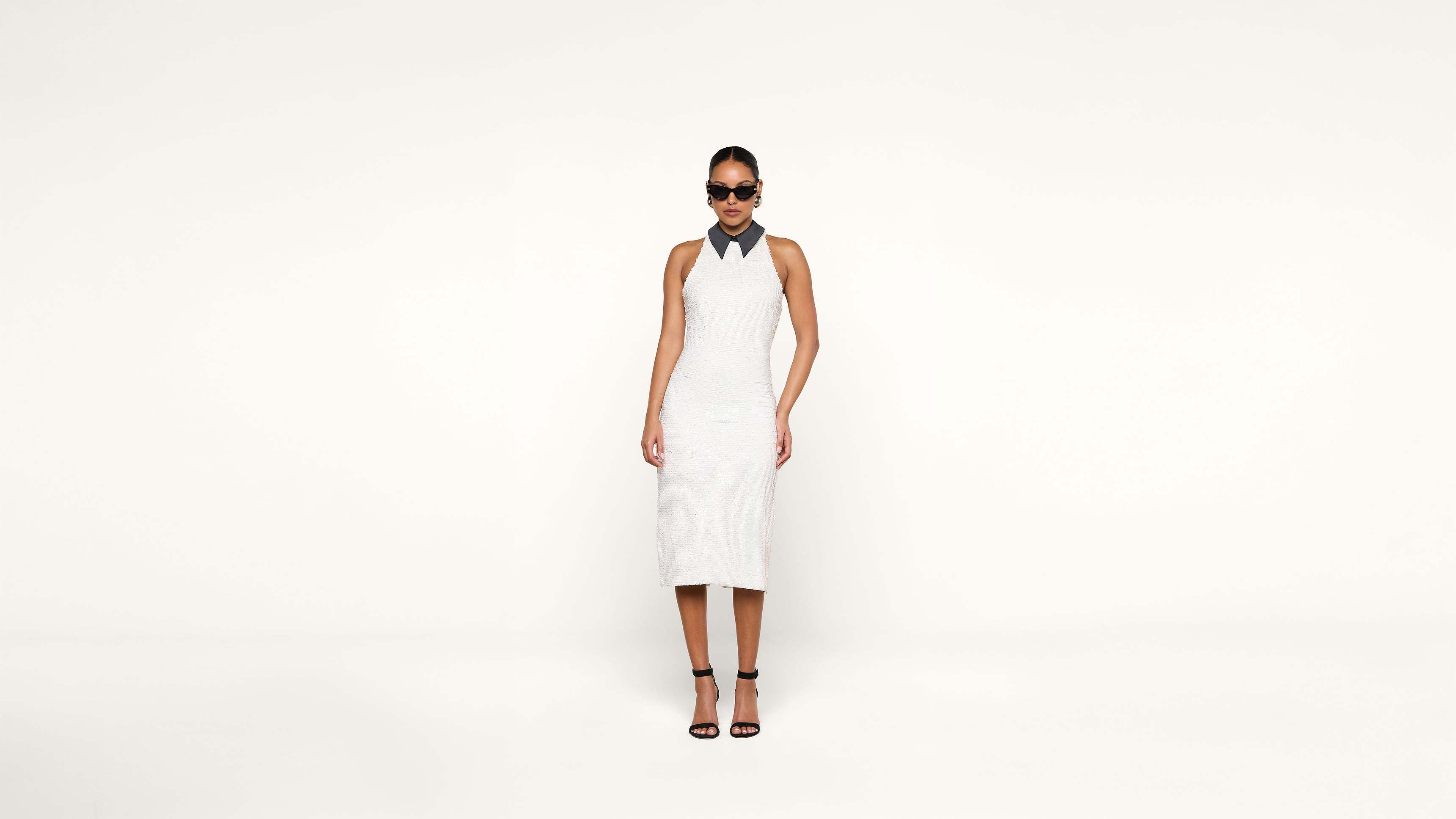 A woman wearing a sleeveless white dress, black strappy heels, and dark sunglasses stands against a plain white background.