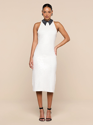 A woman faces forward against a beige background in the Lilith Sequin Dress, a sleeveless, knee-length white party dress with a black poplin collar. She pairs it with black ankle-strap heels and hoop earrings, one hand resting on her hip.