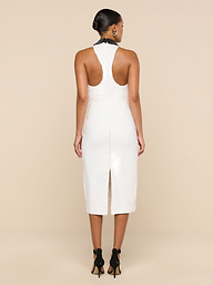 A woman stands facing away, wearing the Lilith Sequin Dress—a fitted, sleeveless party dress in white that falls to the knee and features a back slit. Her dark hair is styled in a low bun, and she pairs the look with black high heels against a plain, light-colored background.