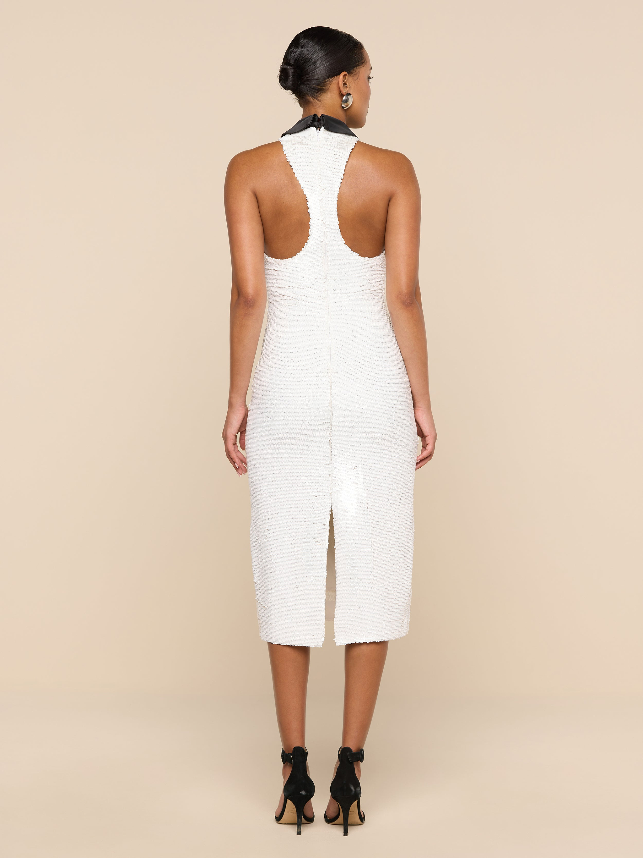 A woman stands facing away, wearing the Lilith Sequin Dress—a fitted, sleeveless party dress in white that falls to the knee and features a back slit. Her dark hair is styled in a low bun, and she pairs the look with black high heels against a plain, light-colored background.