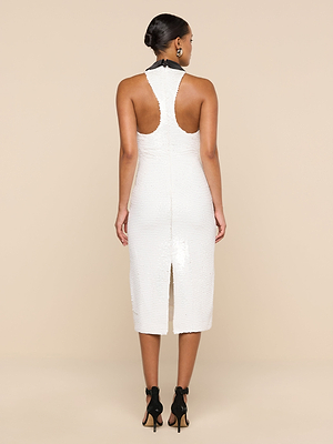 A woman faces away in the Lilith Sequin Dress—white, sleeveless, knee-length with a racerback and back slit. Her hair is in a bun, she wears black ankle-strap high heels, and the backdrop is beige.