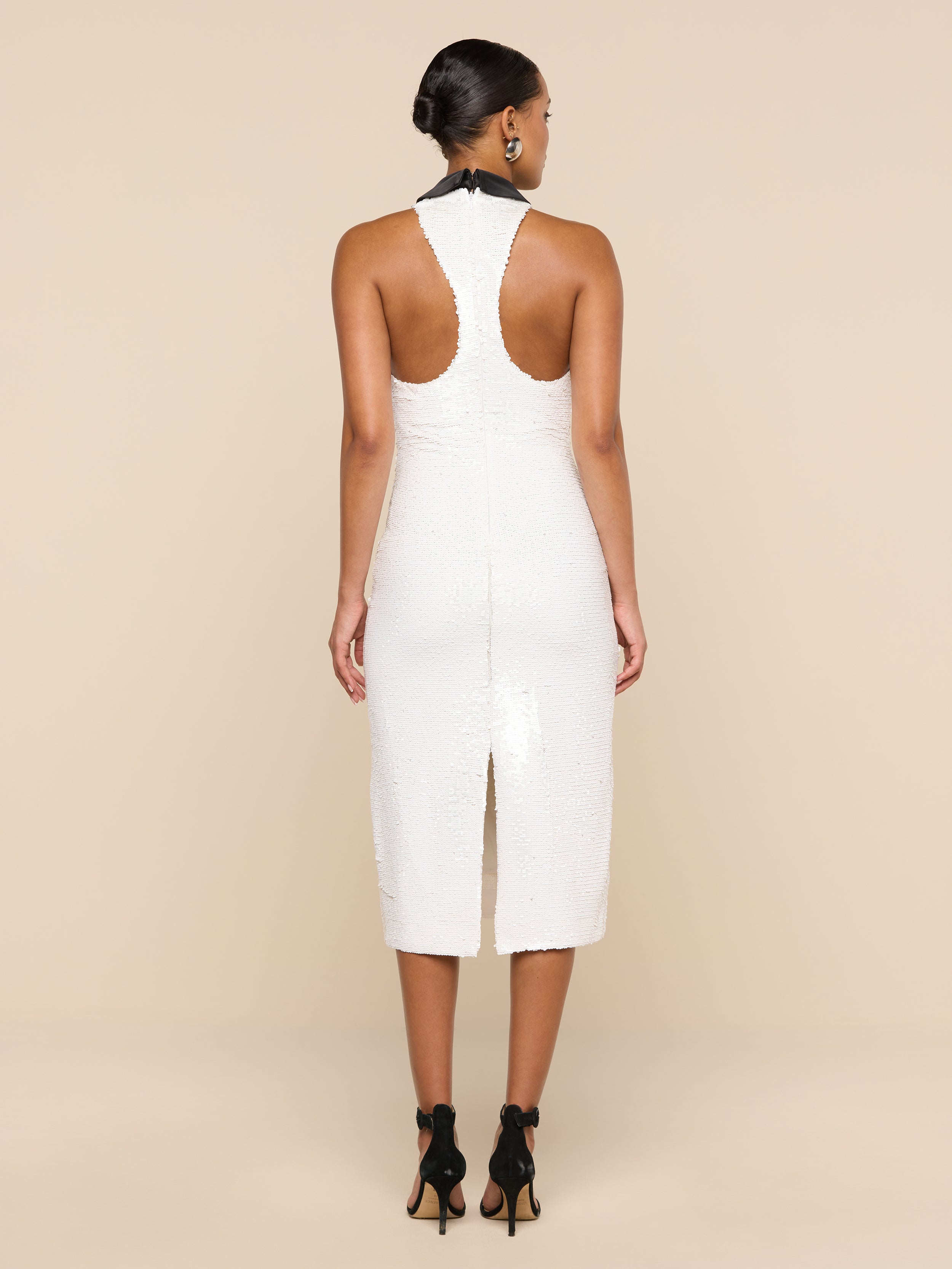 A woman stands facing away, wearing the Lilith Sequin Dress—a fitted, sleeveless party dress in white that falls to the knee and features a back slit. Her dark hair is styled in a low bun, and she pairs the look with black high heels against a plain, light-colored background.