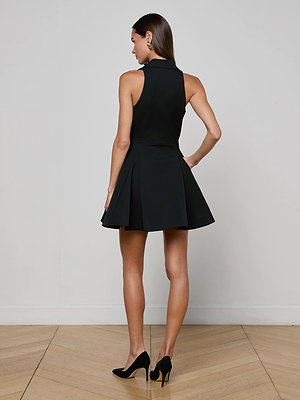 A woman faces away from the camera on a wooden floor, wearing the L'AGENCE Royalty Mini Dress—sleeveless, black, fit-and-flare with a flared skirt—and black high heels against a plain light-colored wall.