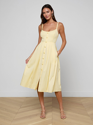 A woman poses indoors wearing the Reanne Midi Dress, a sleeveless pale yellow poplin midi with spaghetti straps, smocked back, button front, and pleated skirt. She pairs it with clear heeled sandals on a light wood floor against a gray wall.