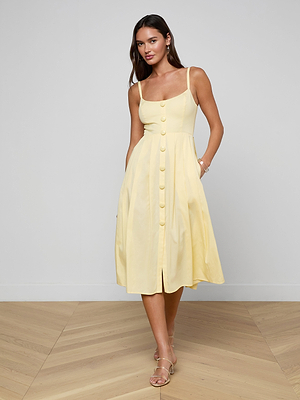 A woman stands on a wooden floor against a light gray wall, wearing the Reanne Midi Dress—a sleeveless, light yellow poplin dress with a fitted bodice, smocked back, large front buttons—and beige strappy sandals.