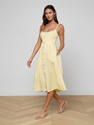 A woman poses with one hand in her pocket, wearing the Reanne Midi Dress—a light yellow poplin midi with large buttons and a smocked back—and strappy heels, standing on a wooden floor against a plain light-colored wall.