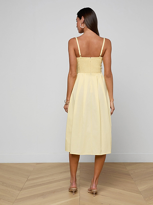 A woman with long brown hair and hoop earrings stands indoors on a wooden floor, seen from behind, wearing the Reanne Midi Dress—a pale yellow midi dress featuring thin straps, a smocked back, and a ruched fitted bodice.