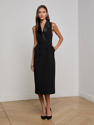A woman stands indoors against a white wall, wearing the L'AGENCE Catherine Tuxedo Midi Dress with a satin lapel and black heels. She has long, straight hair and one hand in her pocket.