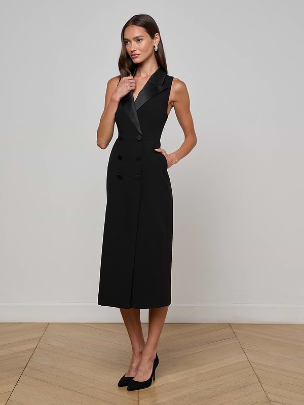 A woman poses in a minimalist room wearing the Catherine Tuxedo Midi Dress by L’AGENCE, featuring a sleeveless design, satin lapel, and notched collar. She pairs it with black heels and casually rests one hand by her face.