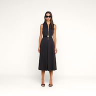 Dressed in the Kolton Dress—a sleek black sleeveless collared midi style with a center zipper and an adjustable gold-buckled belt—a woman pairs her look with black high heels and dark sunglasses while posing against a plain beige background.