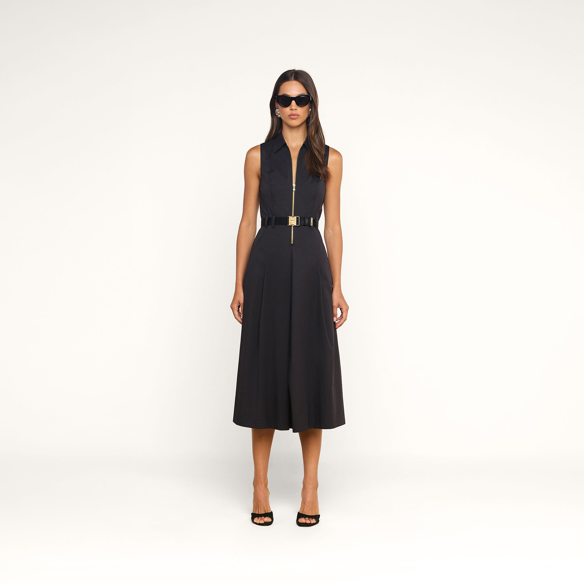 Dressed in the Kolton Dress—a sleek black sleeveless collared midi style with a center zipper and an adjustable gold-buckled belt—a woman pairs her look with black high heels and dark sunglasses while posing against a plain beige background.