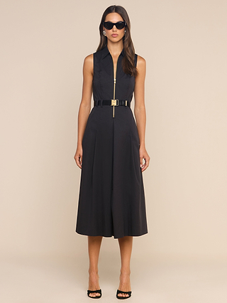 Dressed in the Kolton Dress—a sleek black sleeveless collared midi style with a center zipper and an adjustable gold-buckled belt—a woman pairs her look with black high heels and dark sunglasses while posing against a plain beige background.