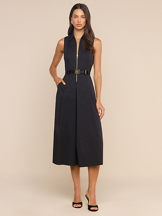 A woman poses against a beige background in the Kolton Dress—a sleeveless black midi dress featuring a center zip and adjustable belt—styled with black open-toe heels.