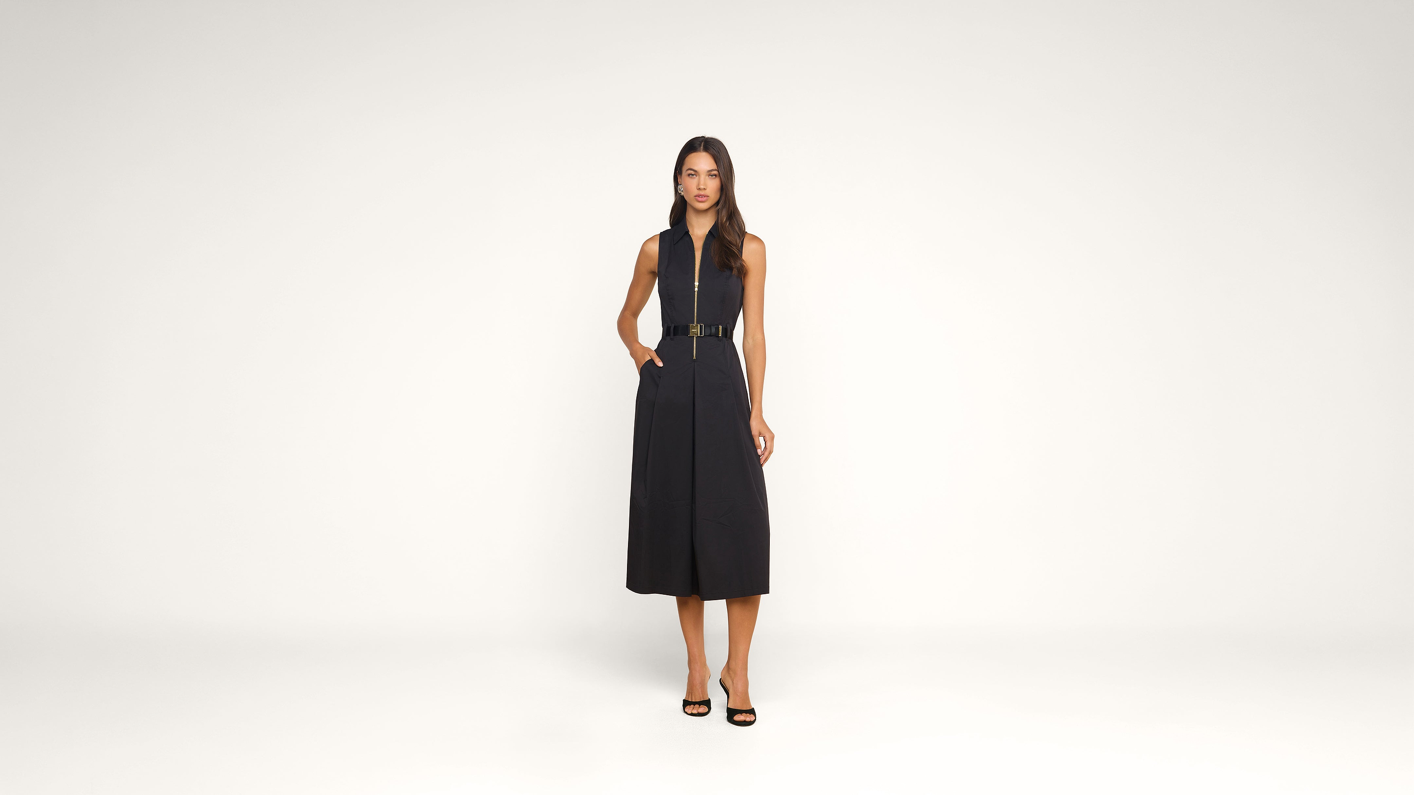 A woman stands in a studio wearing a sleeveless, belted black midi dress and black heels, with her hands in her pockets against a plain white background.