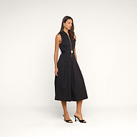 A woman wearing the Kolton Dress—a sleeveless, knee-length black cotton-blend poplin dress featuring a statement belt—and black open-toe heels stands against a plain beige background.