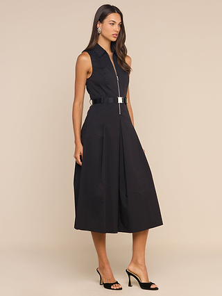 A woman wearing the Kolton Dress—a sleeveless, knee-length black cotton-blend poplin dress featuring a statement belt—and black open-toe heels stands against a plain beige background.