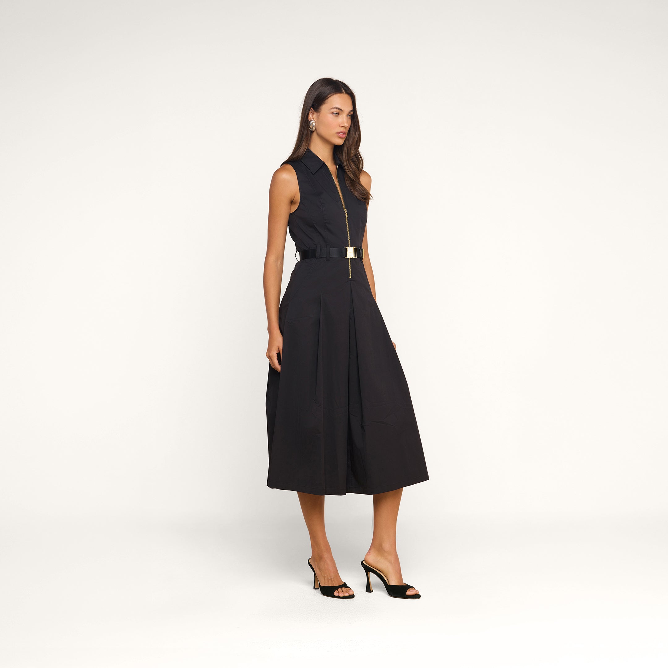 A woman wearing the Kolton Dress—a sleeveless, knee-length black cotton-blend poplin dress featuring a statement belt—and black open-toe heels stands against a plain beige background.