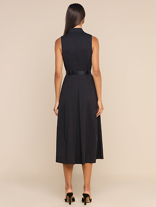A woman stands with her back to the camera, modeling the Kolton Dress—a sleeveless black midi made from cotton-blend poplin—accentuated at the waist with a bold belt and styled with black heeled sandals, set against a simple beige backdrop.