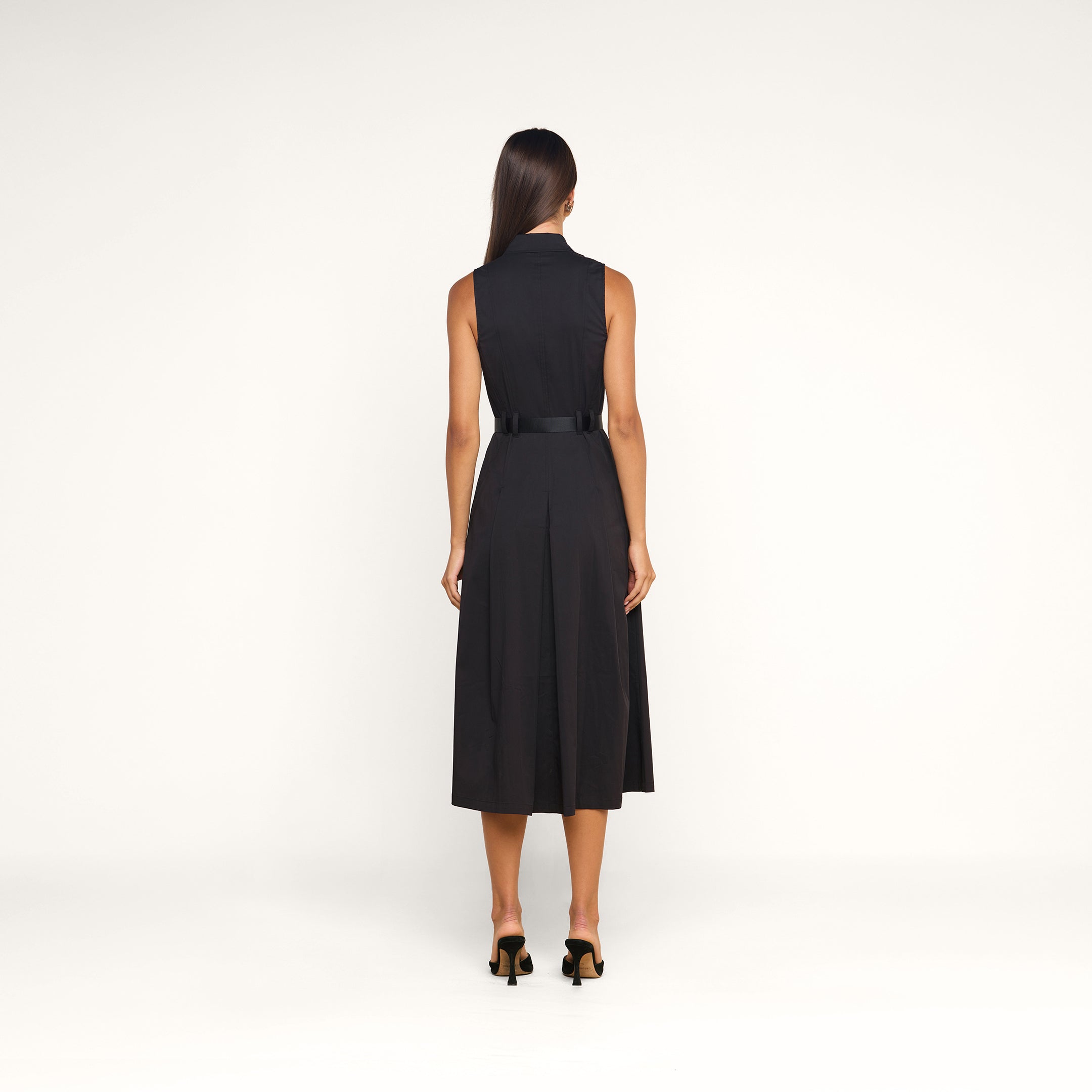A woman stands with her back to the camera, modeling the Kolton Dress—a sleeveless black midi made from cotton-blend poplin—accentuated at the waist with a bold belt and styled with black heeled sandals, set against a simple beige backdrop.