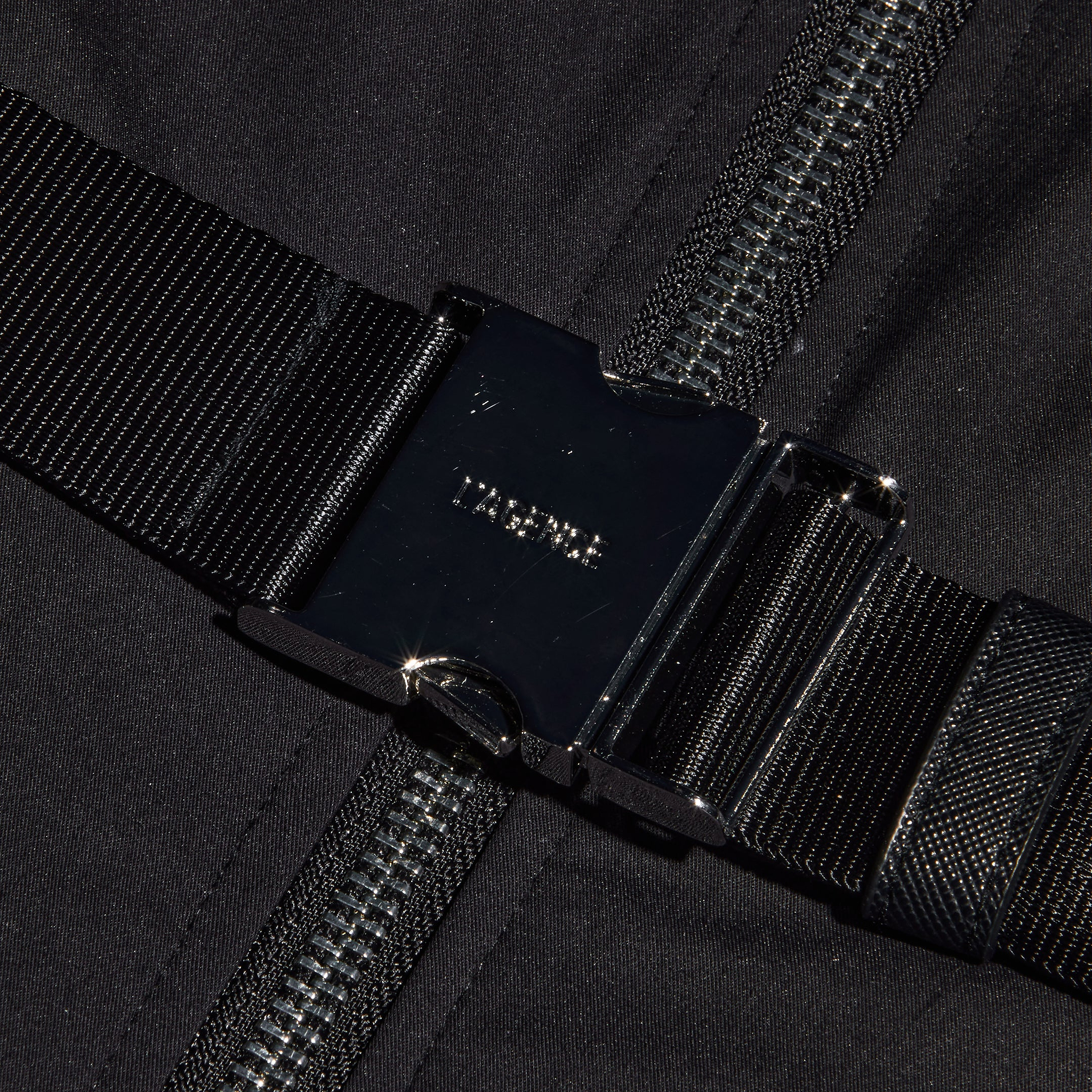 Close-up of a black metal buckle fastened over a dark zipper, with black fabric and webbing straps in the background—bold hardware inspired by statement belt details designed for the Kolton Dress.