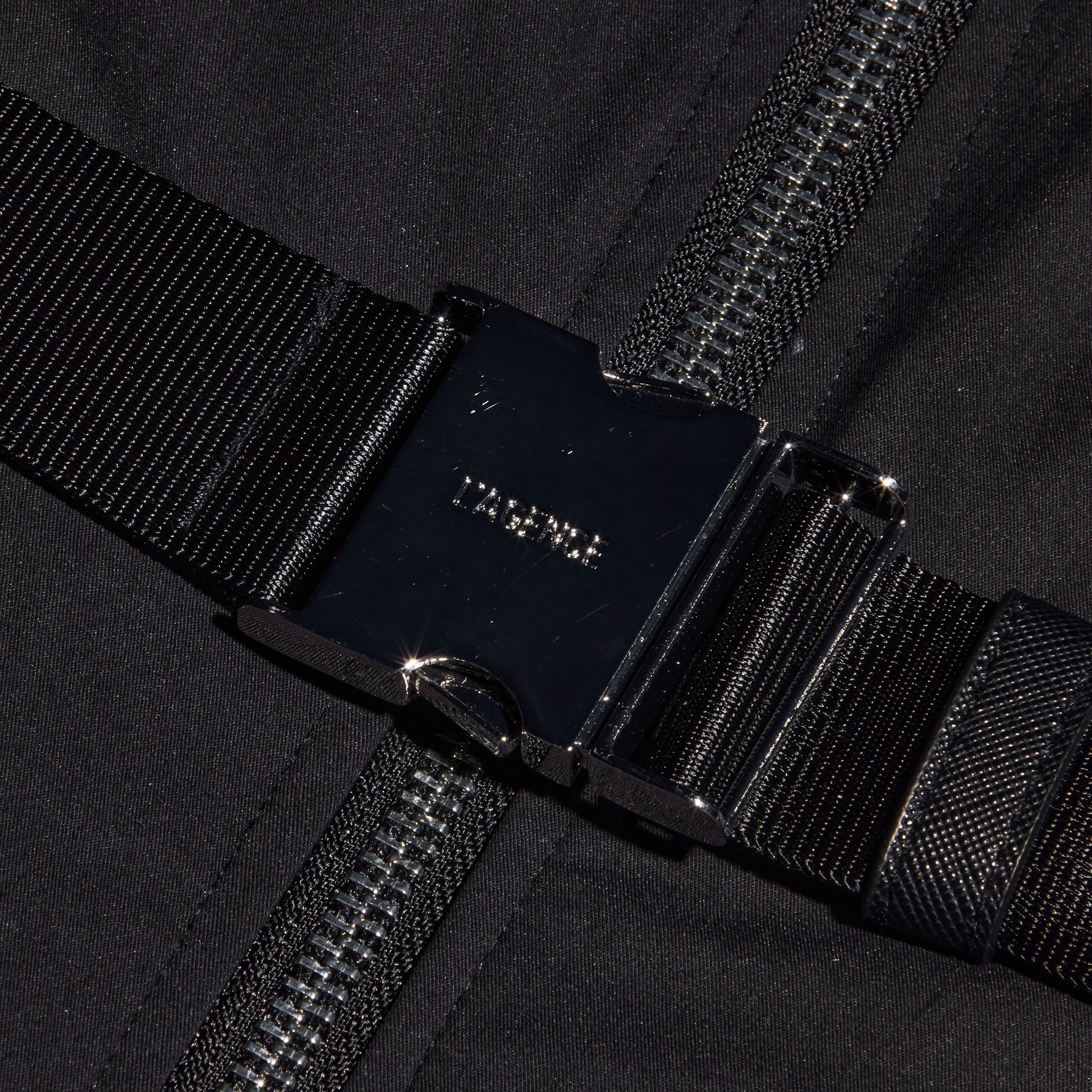 Close-up of a black metal buckle fastened over a dark zipper, with black fabric and webbing straps in the background—bold hardware inspired by statement belt details designed for the Kolton Dress.