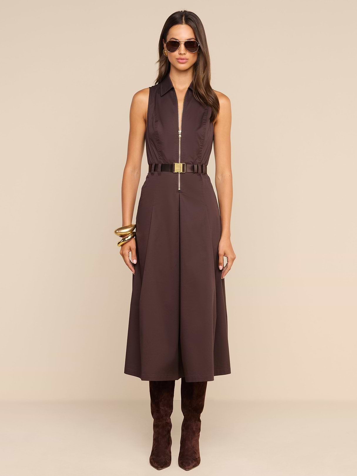 A woman wearing the Kolton Dress—a sleeveless brown cotton-blend poplin midi dress—styled with a gold belt, large bangles, sunglasses, and knee-high brown boots stands against a plain white background.