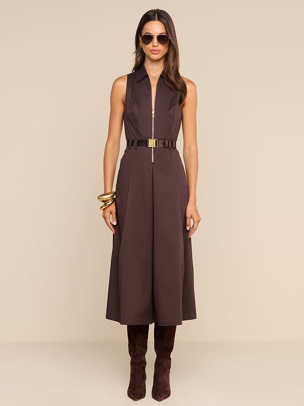 A woman wearing the Kolton Dress—a sleeveless brown cotton-blend poplin midi dress—styled with a gold belt, large bangles, sunglasses, and knee-high brown boots stands against a plain white background.