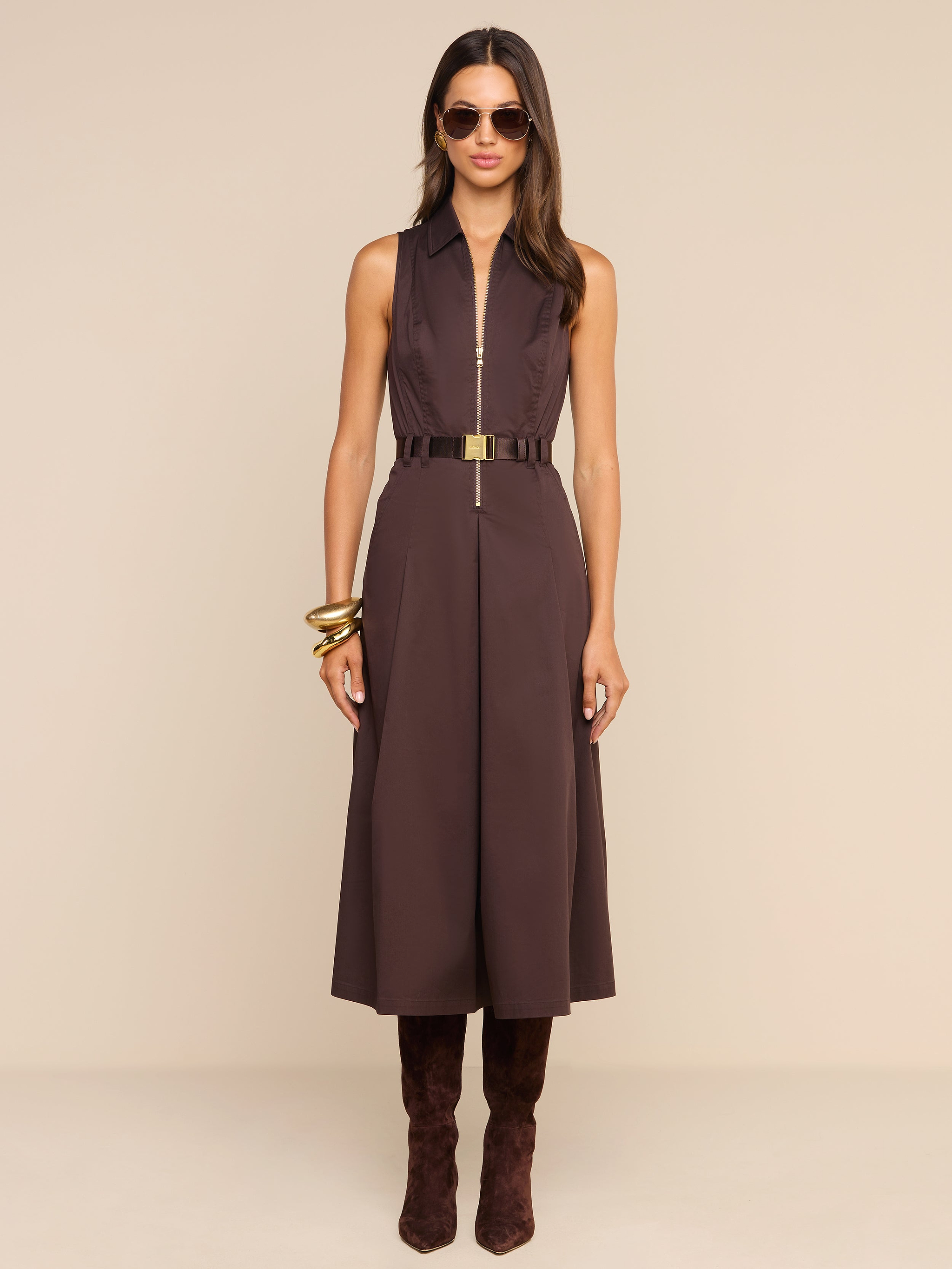A woman wearing the Kolton Dress—a sleeveless brown cotton-blend poplin midi dress—styled with a gold belt, large bangles, sunglasses, and knee-high brown boots stands against a plain white background.